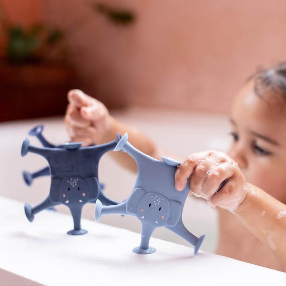 A toddler in a bathtub plays with two blue elephant-shaped bath toys with suction feet. The scene is playful and focused on bathtime fun.