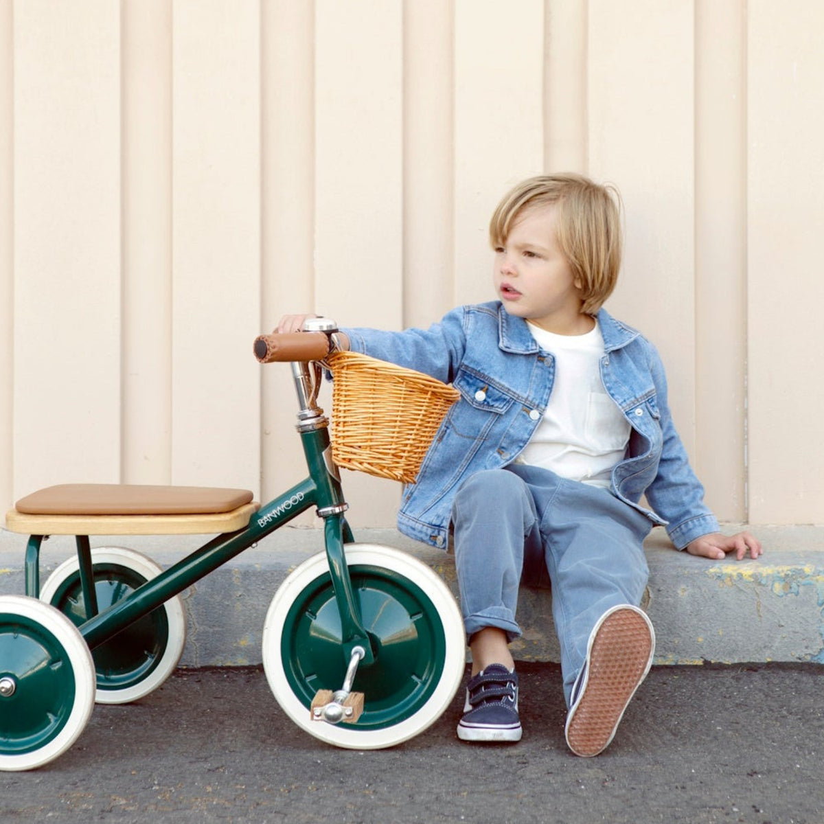 Young child in denim pushing a green vintage tricycle on a paved sidewalk near a house.