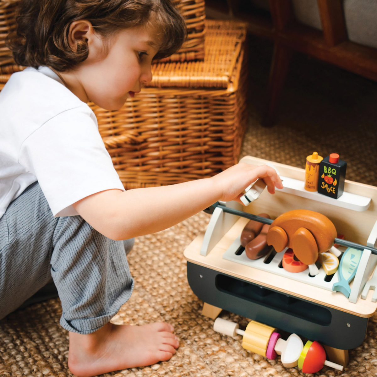 Child reaching to play with a wooden rotisserie chicken on a toy barbecue set.