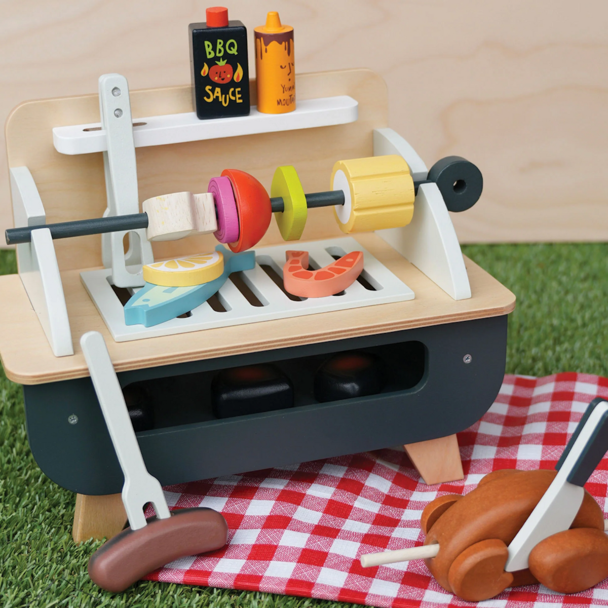 Wooden toy barbecue grill with skewer, play food, and utensils on a red checkered picnic blanket.