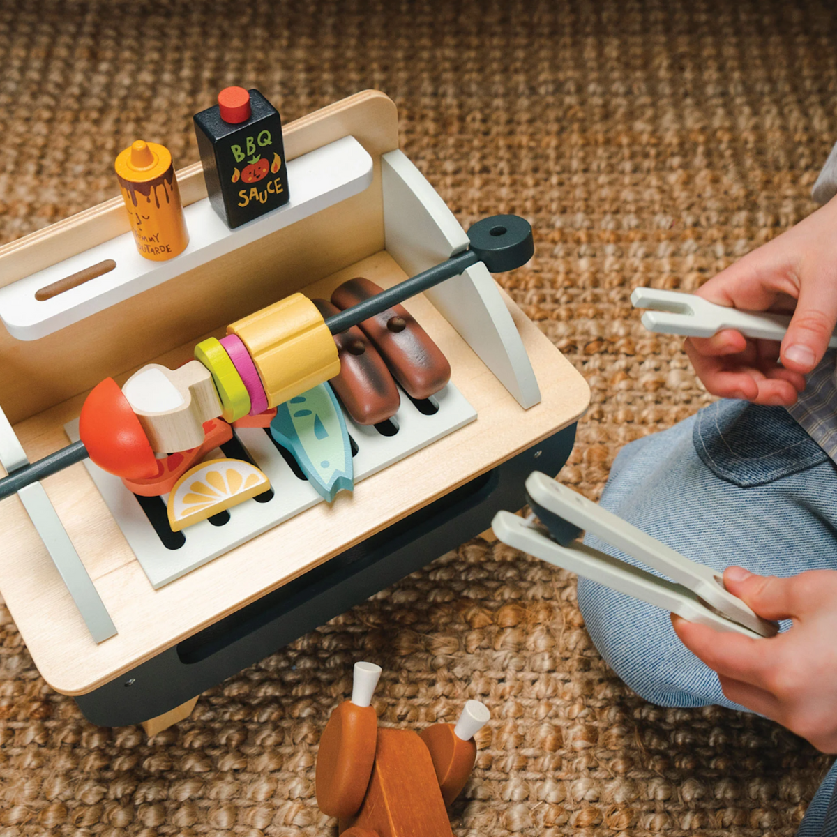 Overhead view of a child using wooden tongs to play with a barbecue set on a woven rug.
