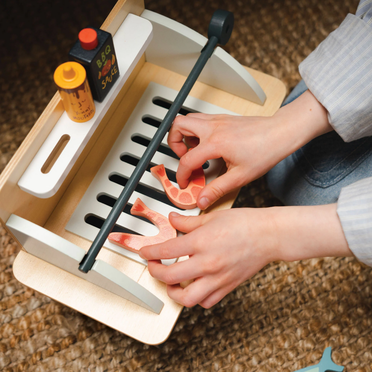 Close-up of hands arranging wooden play prawns on the grill of a toy barbecue set.