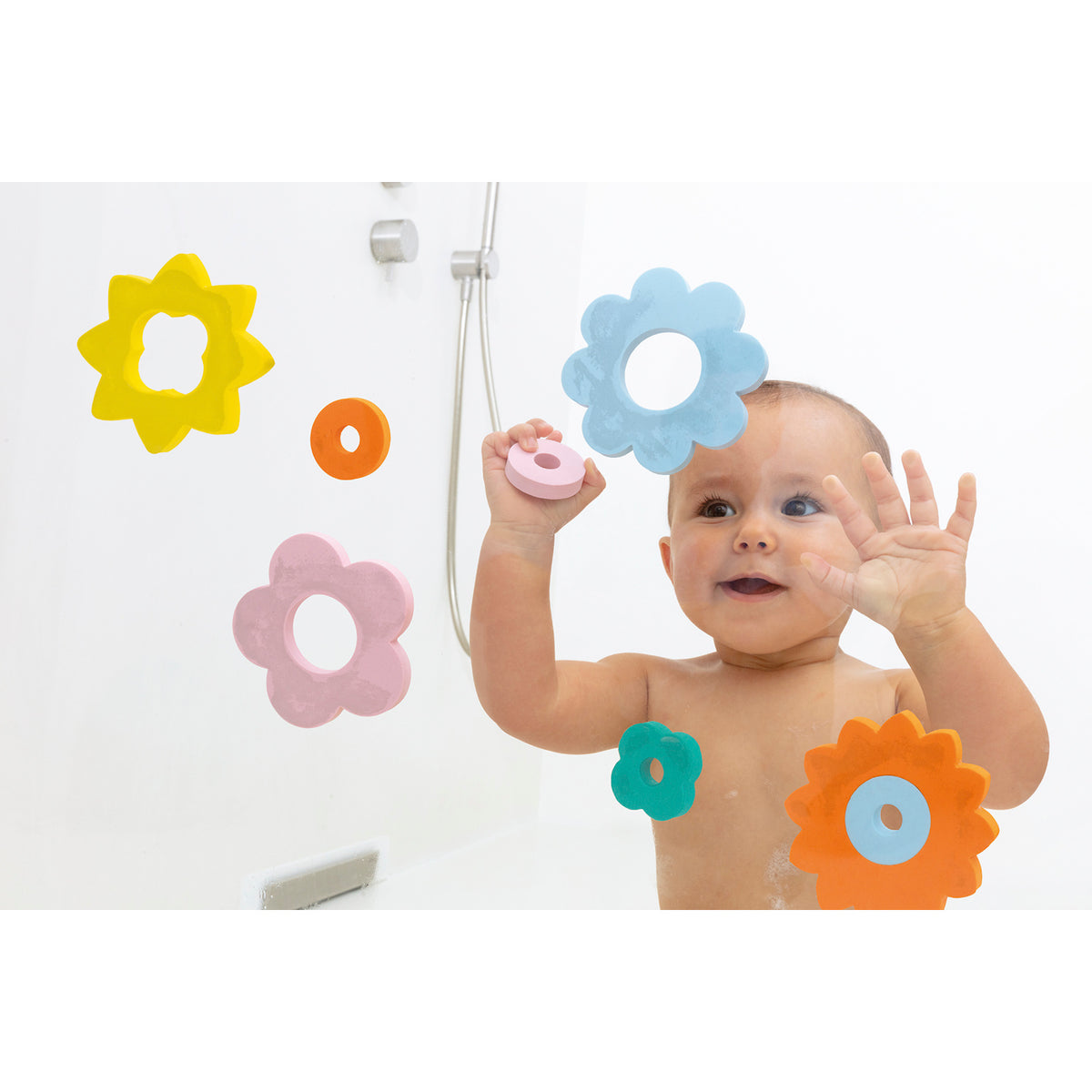 A baby in a bath, smiling and playing with colorful flower-shaped foam toys stuck to a glass surface. The mood is playful and joyful.