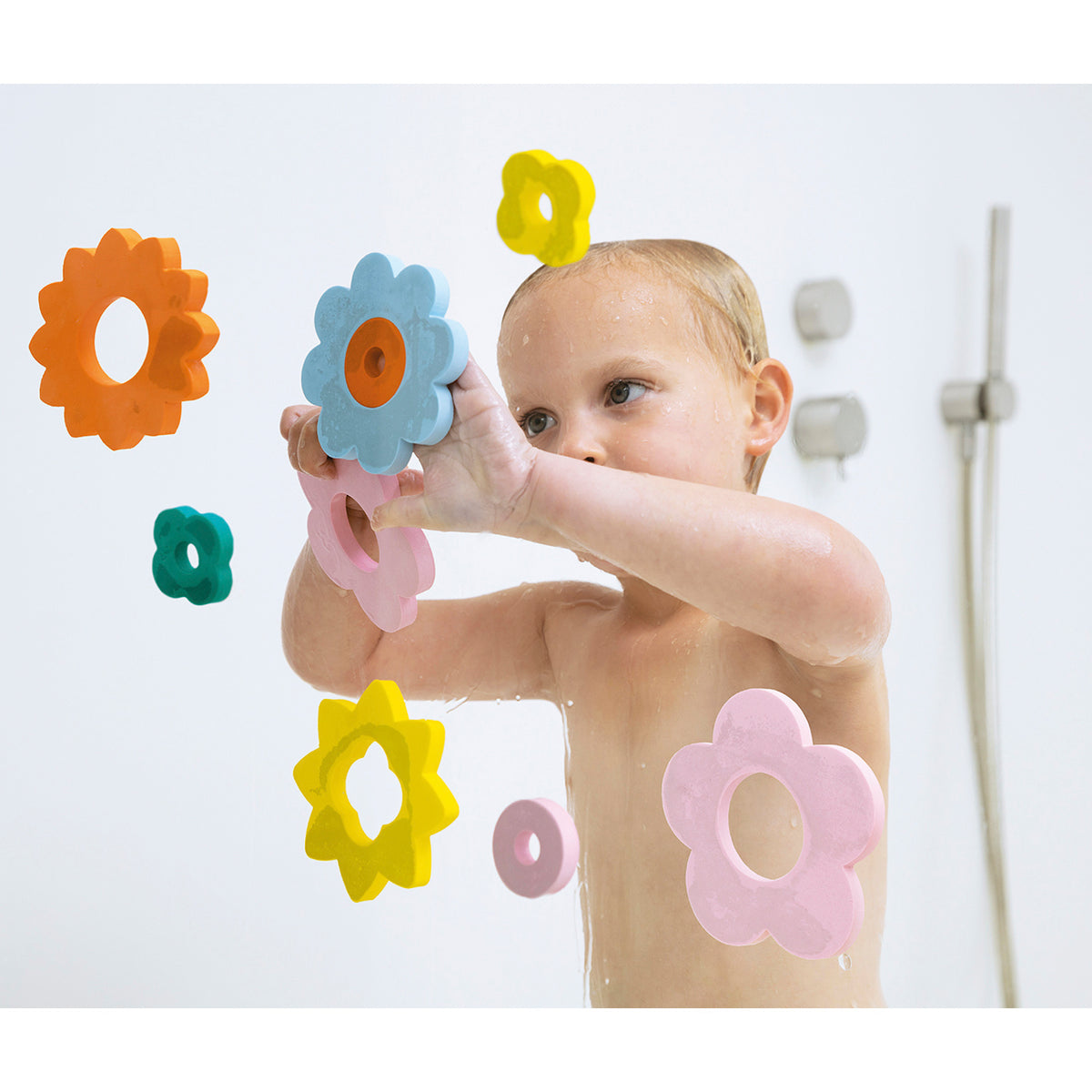 A baby in a bath, smiling and playing with colorful flower-shaped foam toys stuck to a glass surface. The mood is playful and joyful.
