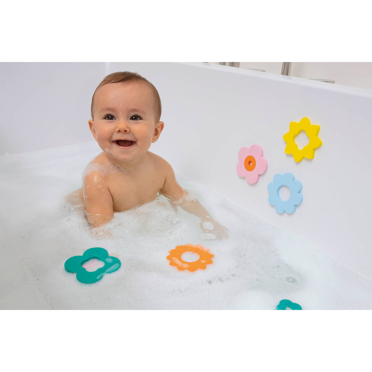 A smiling baby sits in a white bathtub filled with bubbles, surrounded by colorful flower-shaped toys on the wall, creating a playful, joyful atmosphere.
