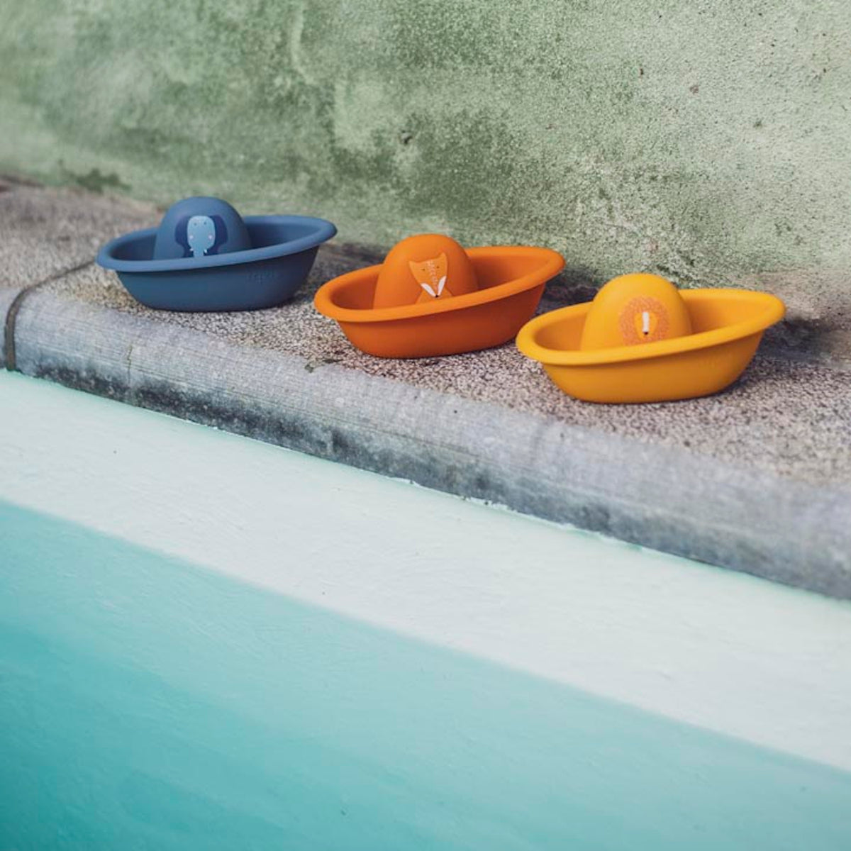 Three colorful toy boats with animal faces lined up on the edge of a pool.