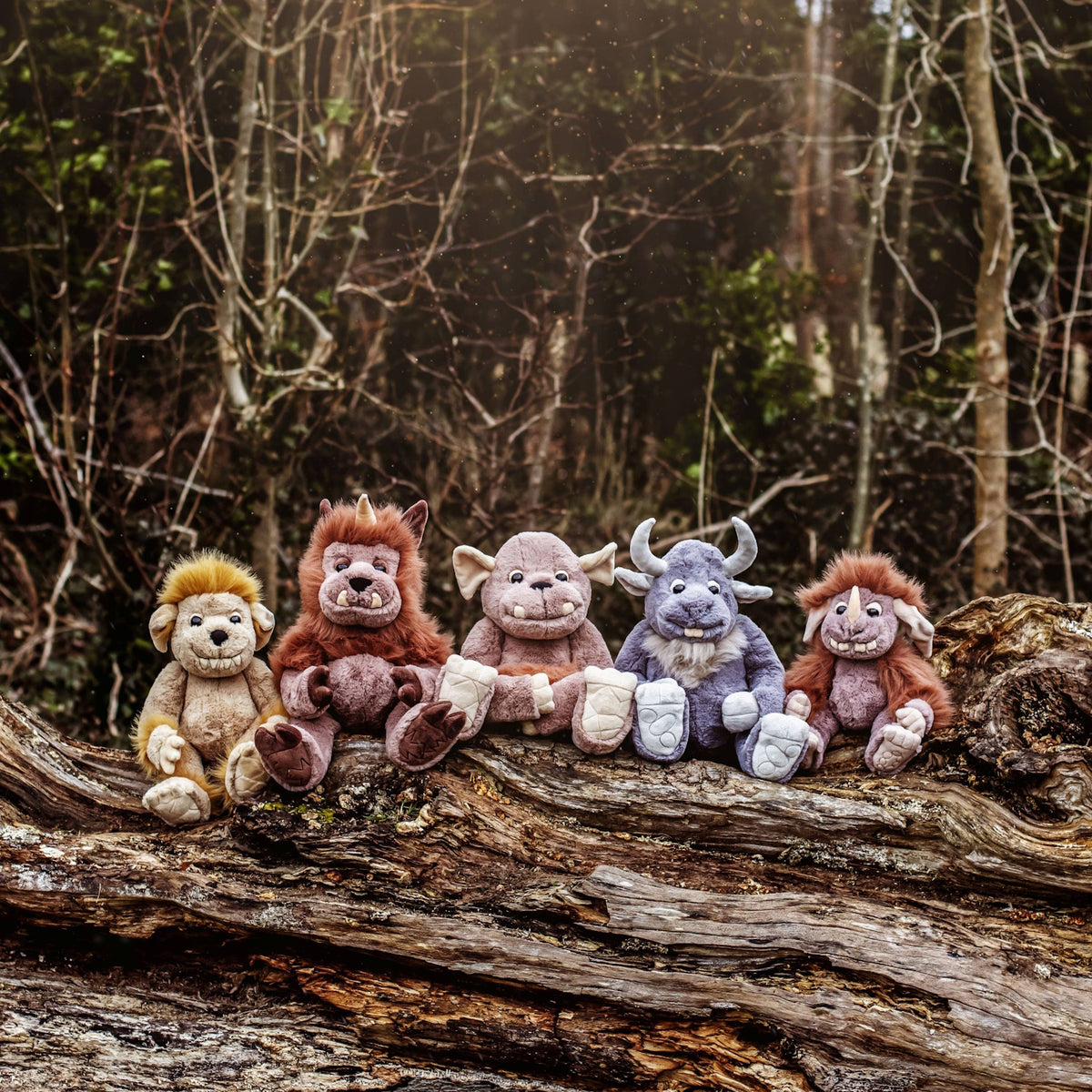 Five plush monster and creature toys lined up sitting in a row on a large fallen tree trunk in a forest.