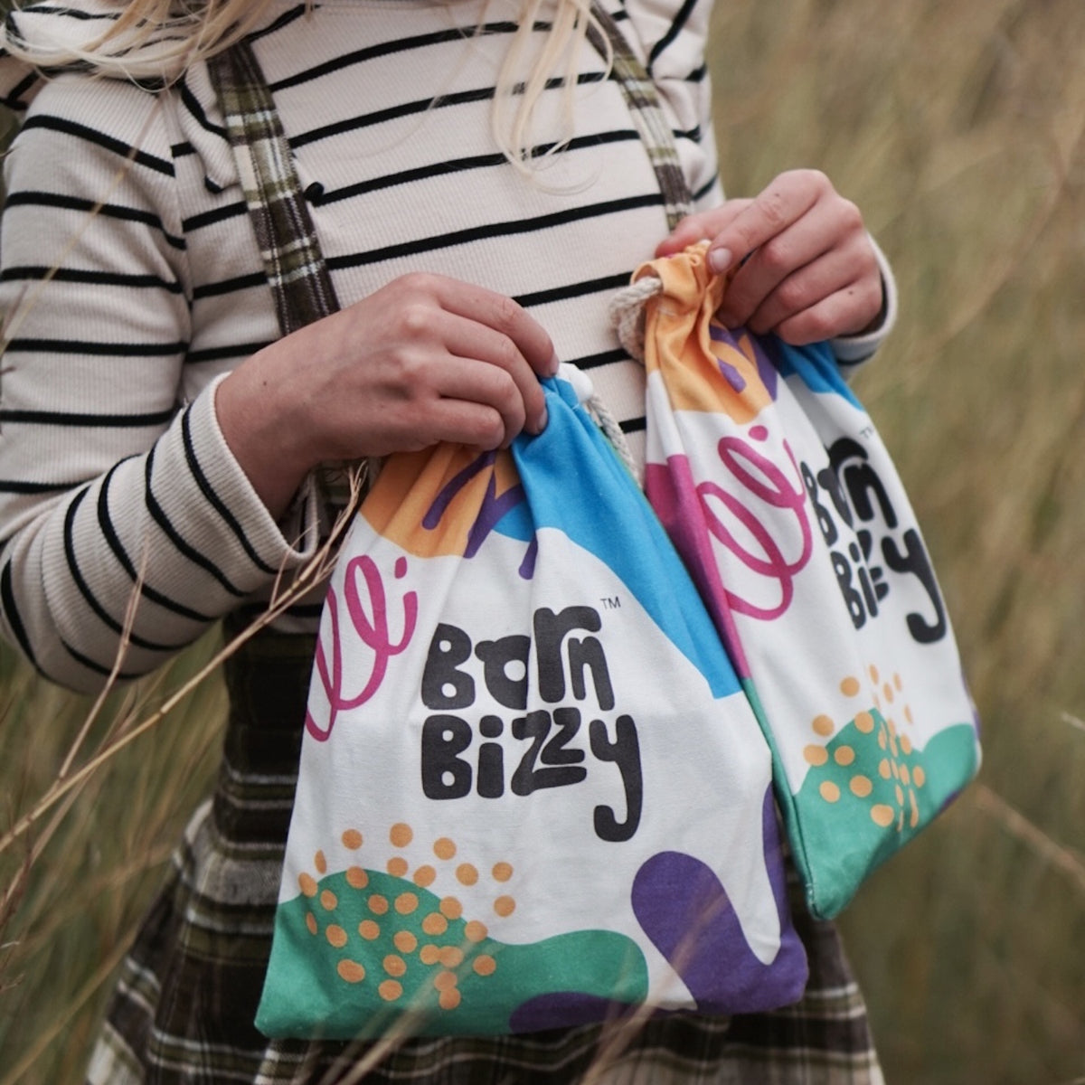 Child holding two colorful drawstring canvas bags with "born bizzy" text printed on them in a grassy field.