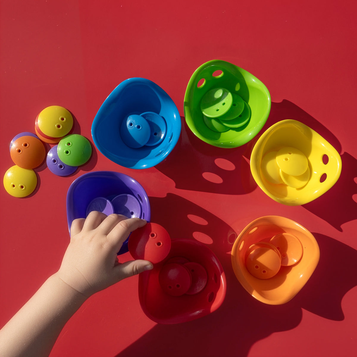 Child's hand sorting colorful plastic toy disks into matching colored bowls on a vibrant red surface.