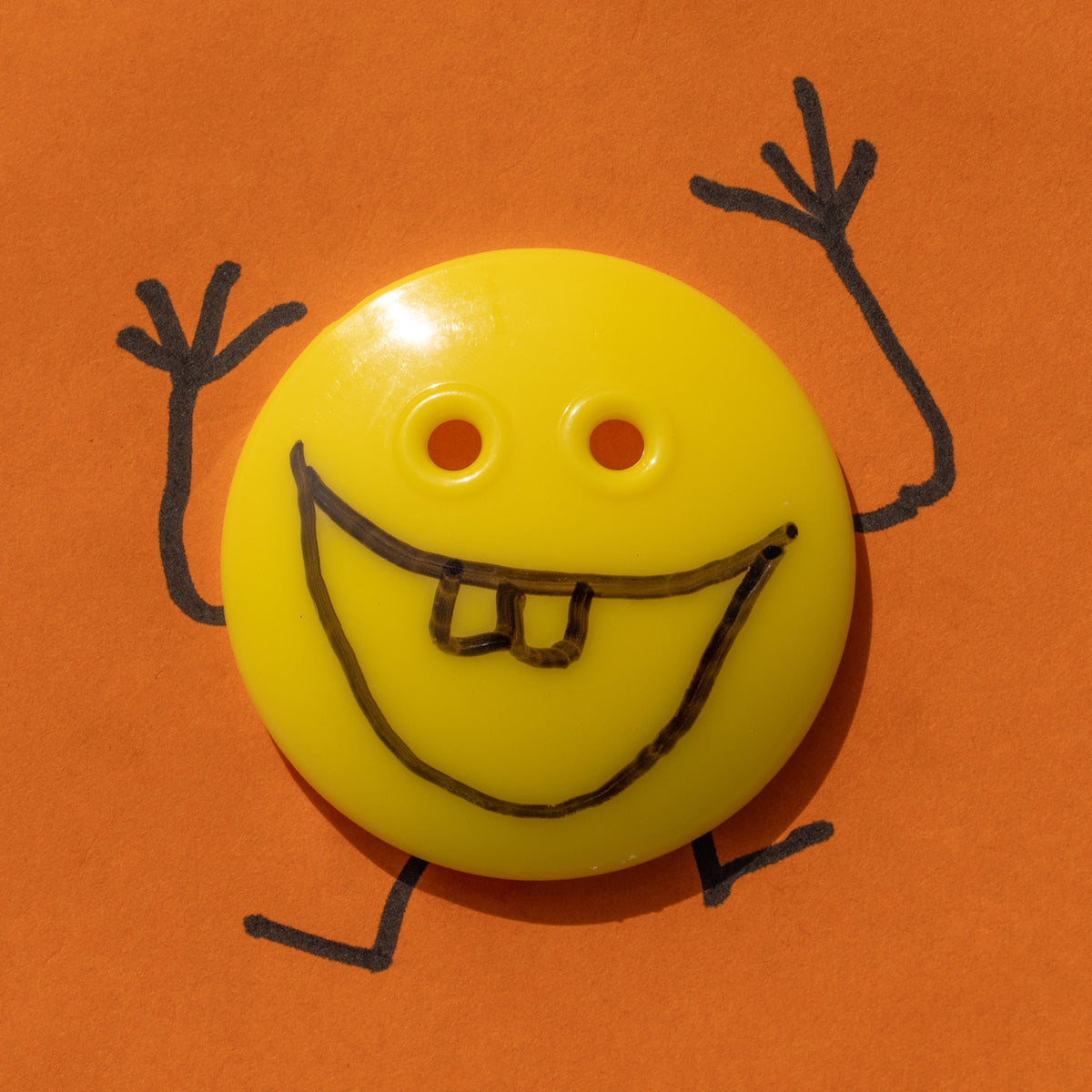A quirky image of a yellow button face with hand-drawn marker limbs and buck teeth, against a bright orange background.