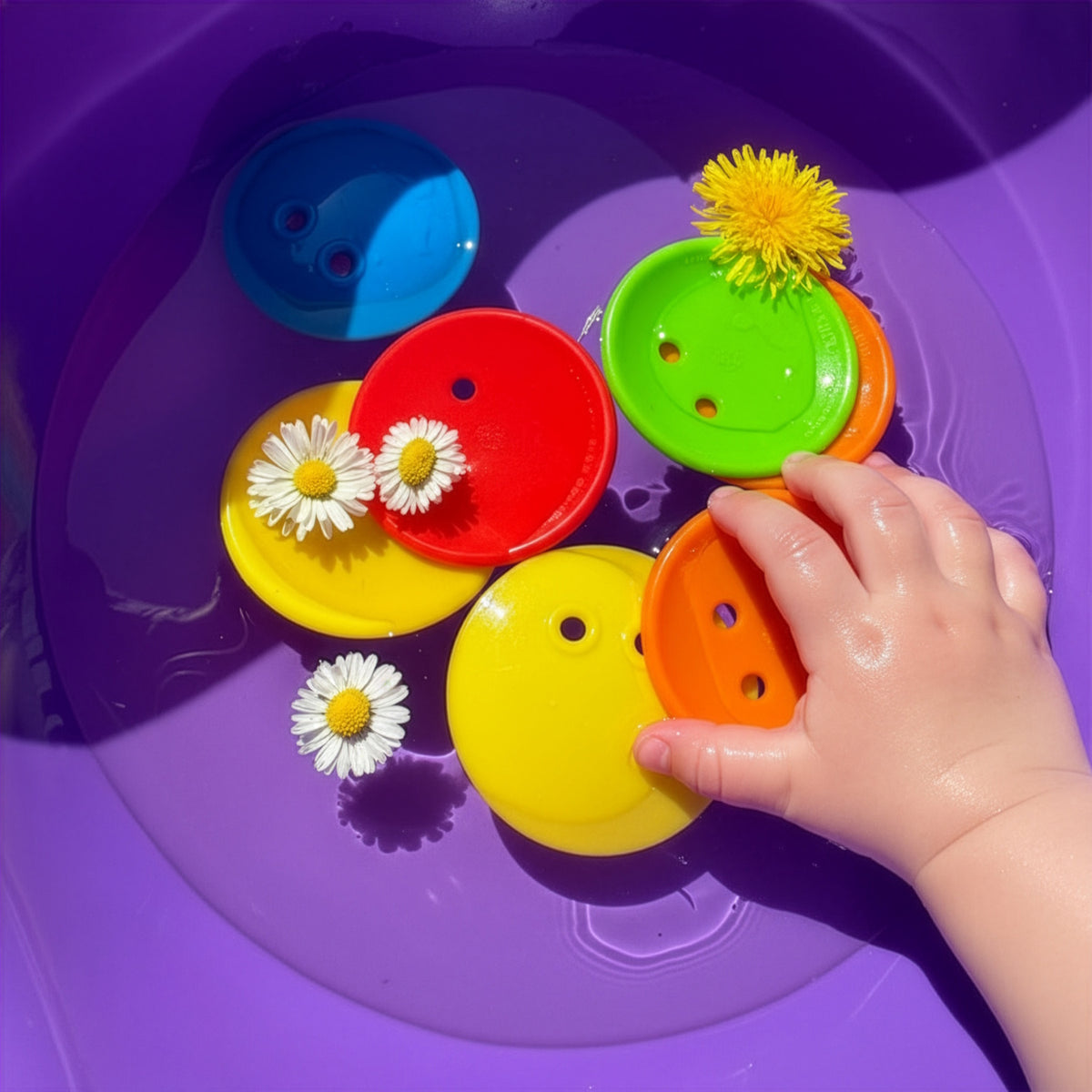A child's hand playing with colorful Bilibo Game Pix and floating daisies in a purple water basin for sensory play.