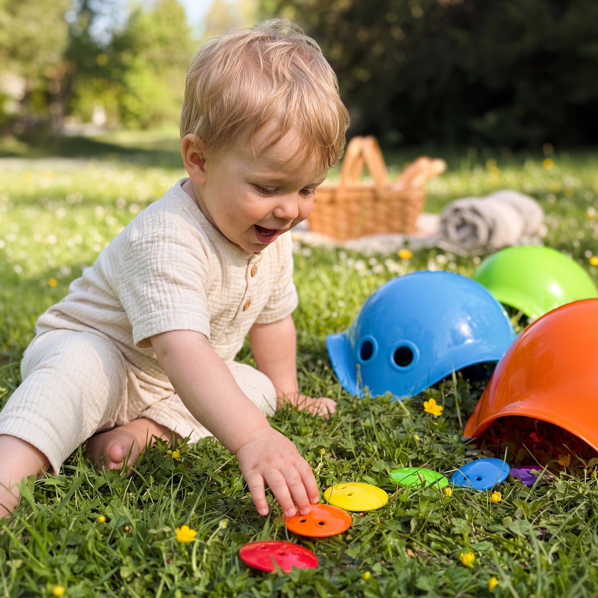 Toddler playing with colorful Bilibo mini shells and toy buttons on green grass during an outdoor picnic.