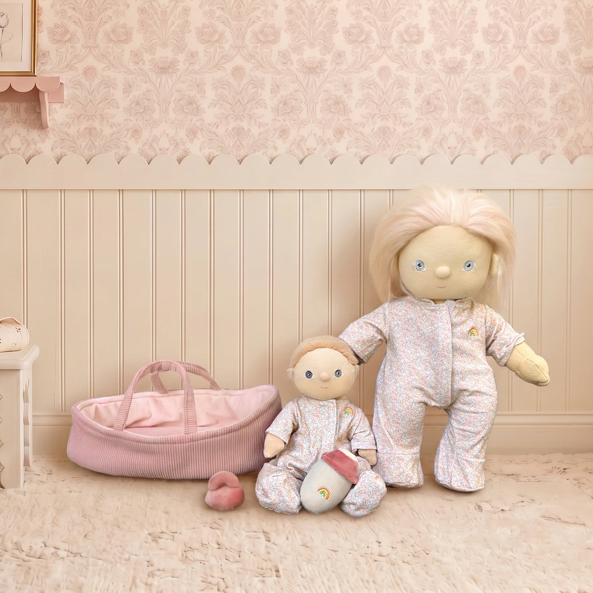 Two Olli Ella Dinkum Dolls in floral pajamas next to a pink bassinet in a vintage nursery.