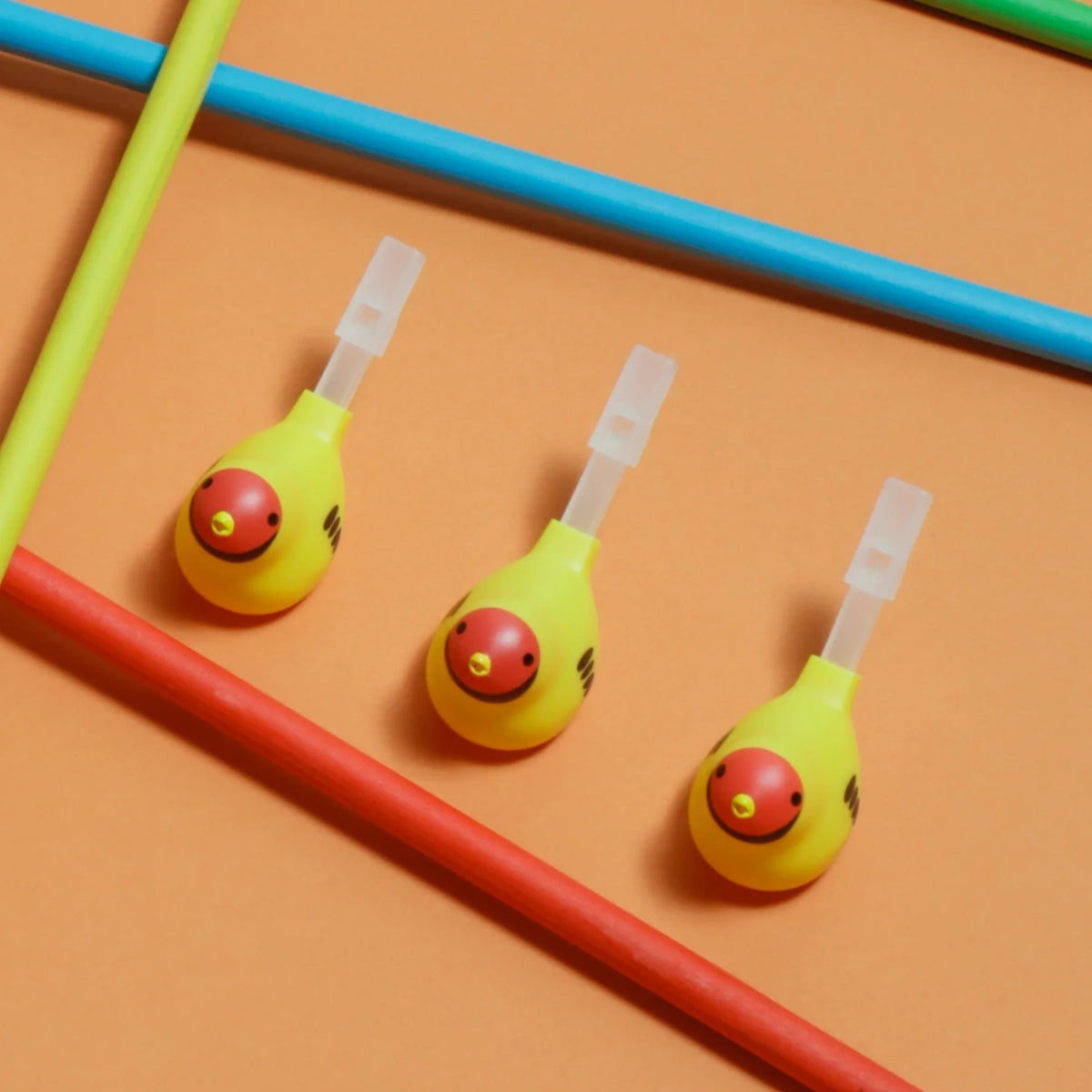 Three yellow bird-shaped whistles with red beaks on an orange background, interspersed with diagonally placed blue, red, and green sticks. Bright and playful tone.