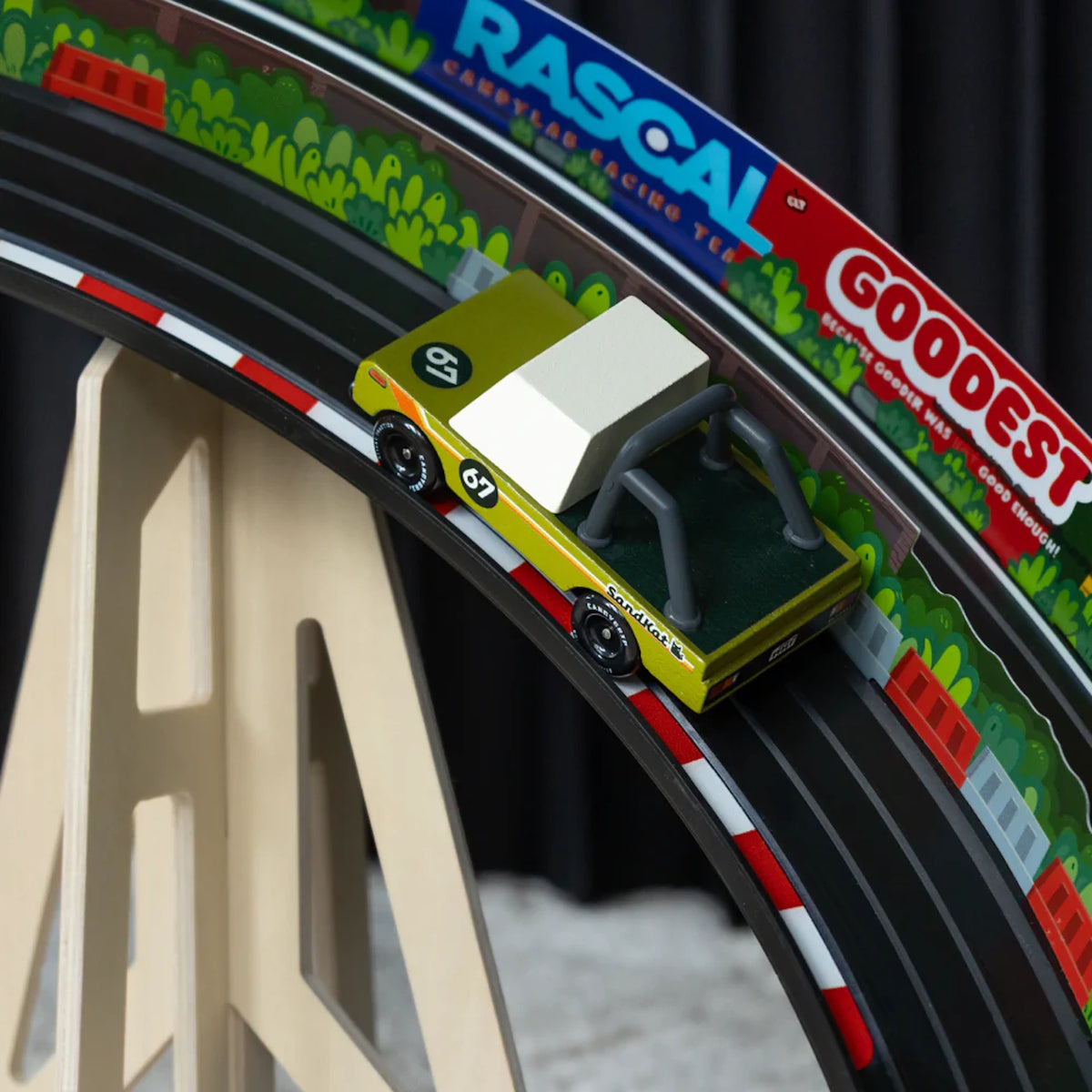 Olive green Sand Kat wooden toy truck racing on a curved Candycar track with colorful Rascal and Goodest racing banners.