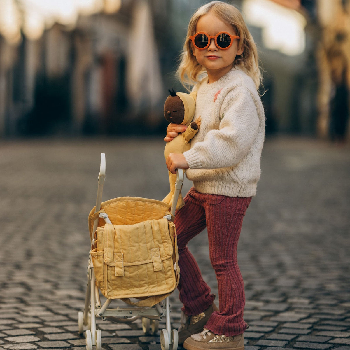 Young girl with sunglasses holding a doll and standing by a yellow Carrie Convertible Changing Set stroller.