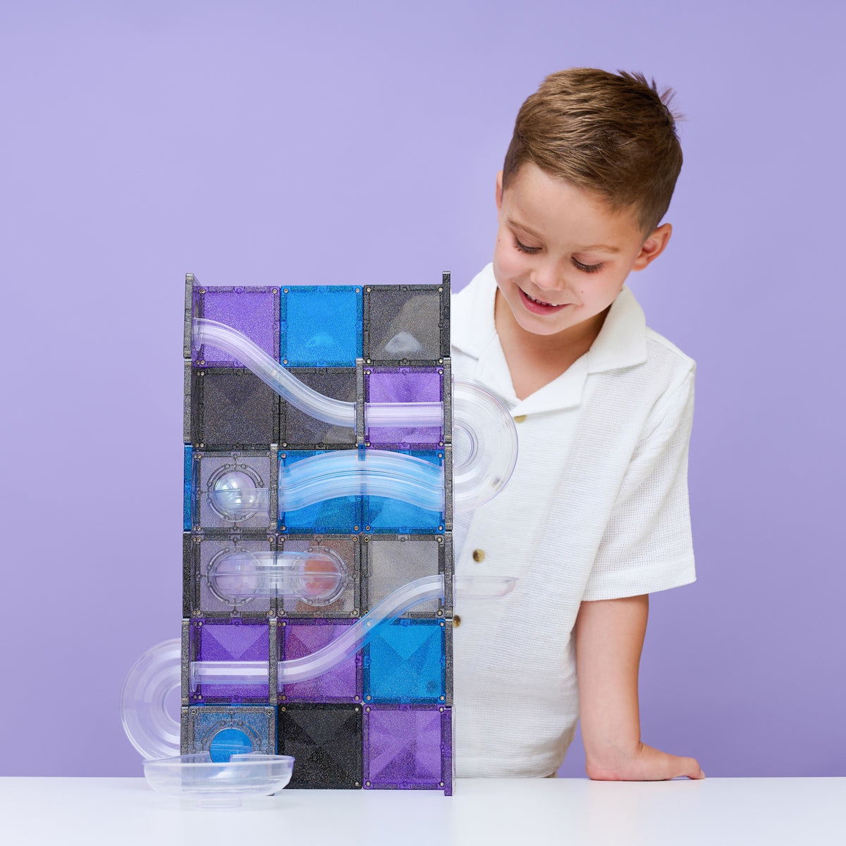 Young boy smiling at a built glittery blue and purple magnetic tile ball run structure.