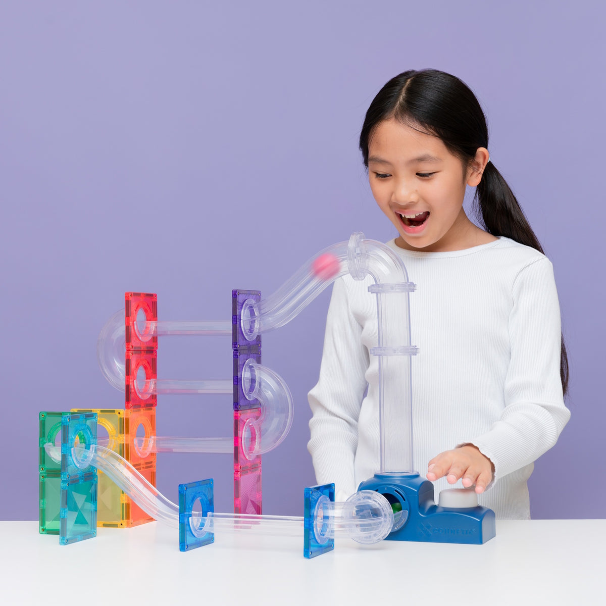 Young girl playing with a colorful magnetic tile ball run structure and launcher on a purple background.