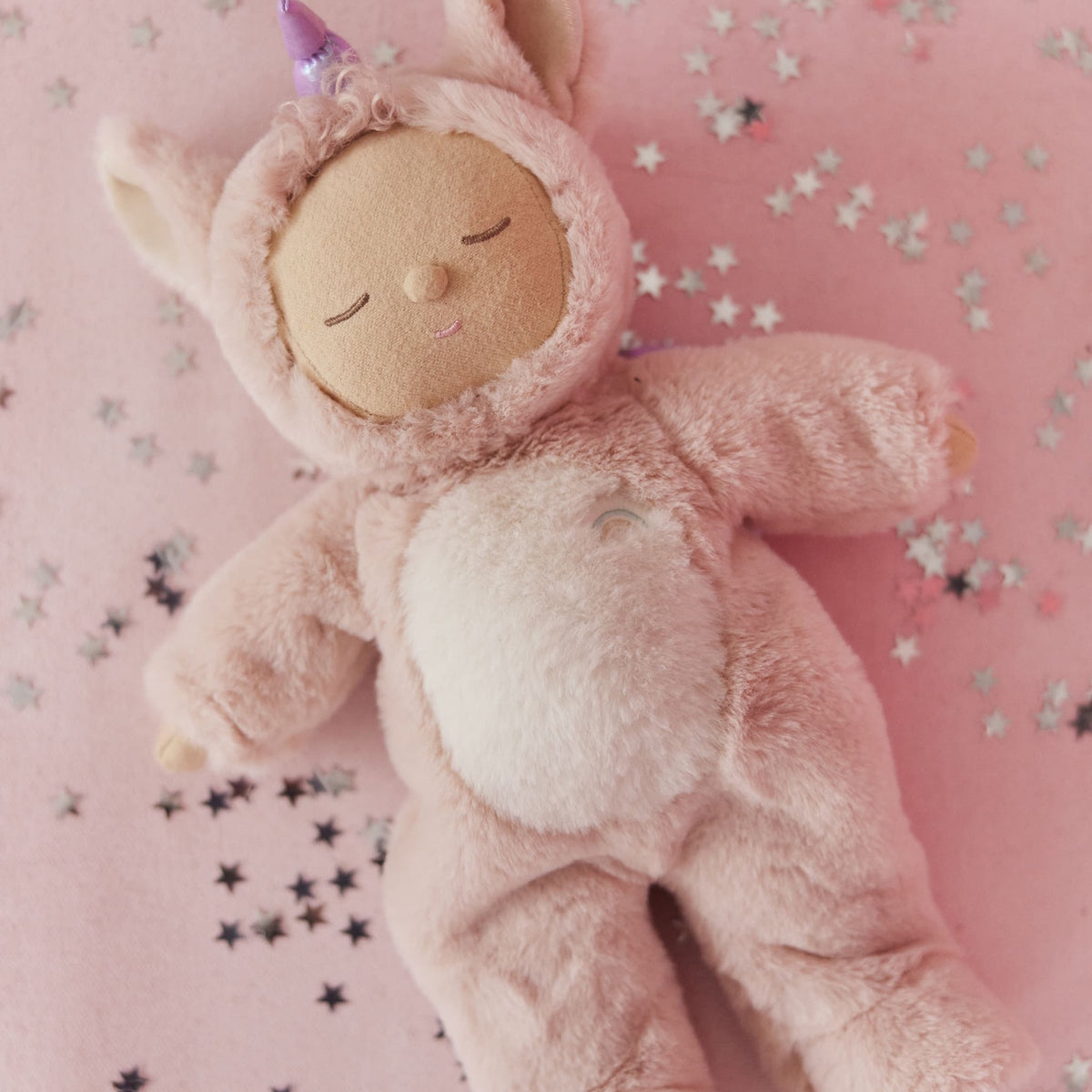 Olli Ella Cozy Dinkum Unicorn Sugarpie plush doll lying on a pink background with star confetti.