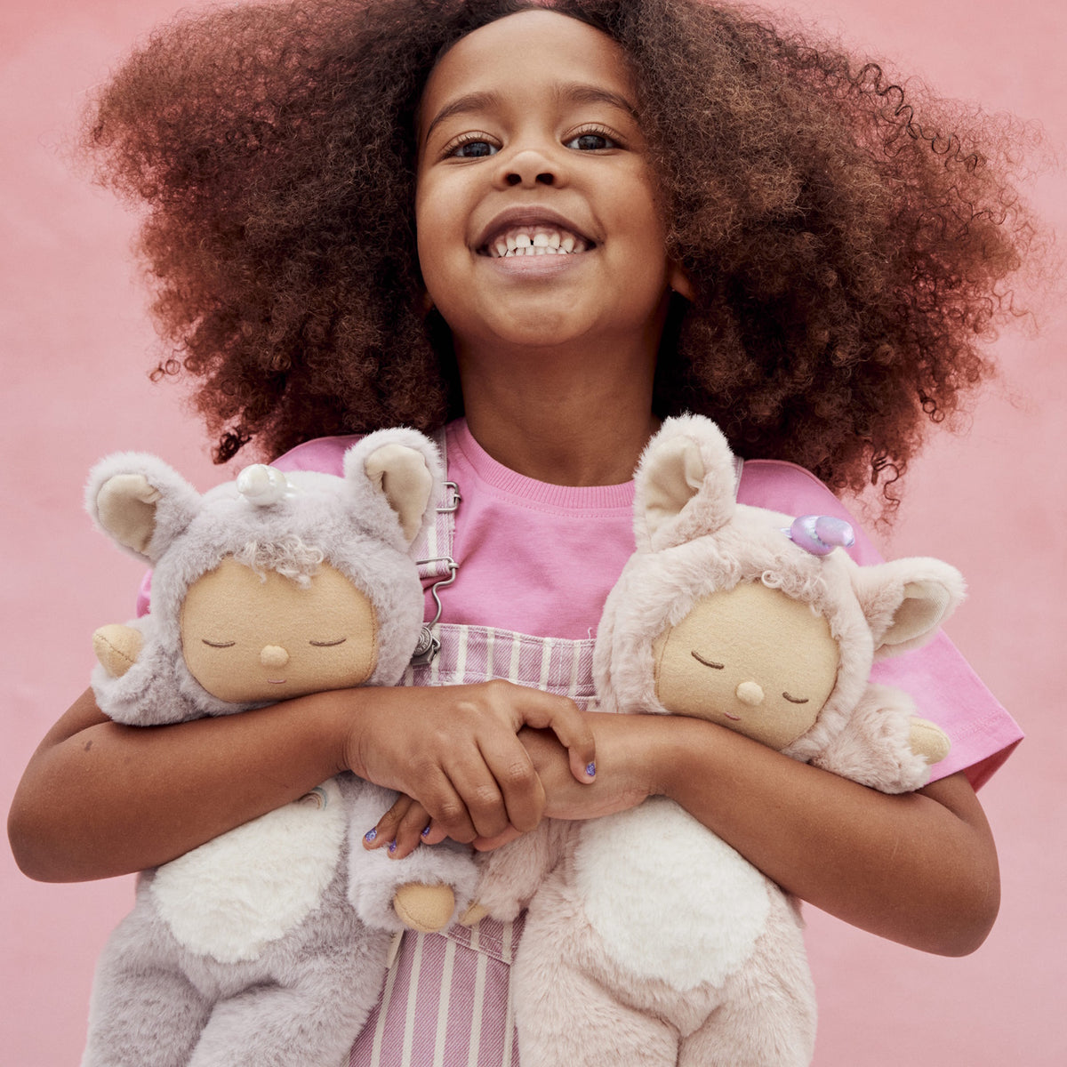 Smiling young girl hugging two plush Olli Ella Cozy Dinkum Dolls against a solid pink background.