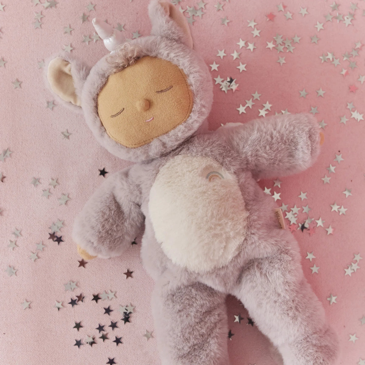Olli Ella Cozy Dinkum Unicorn Sweetpea plush doll lying on a pink starry background.