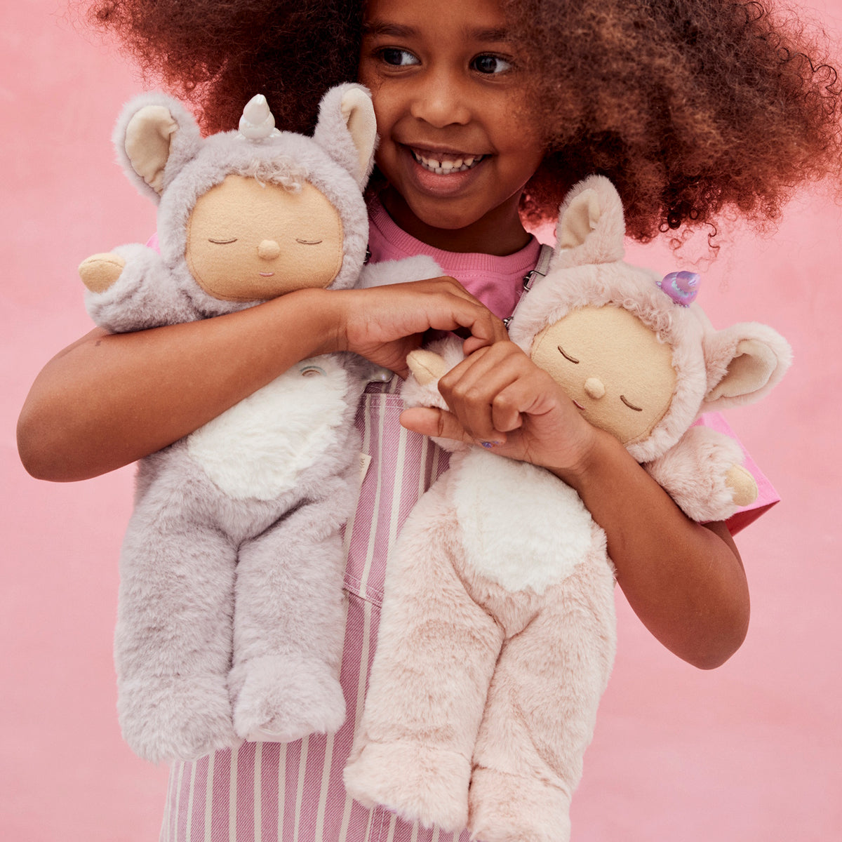 Young girl smiling and holding two Olli Ella Cozy Dinkums plush unicorn dolls against a pink background.