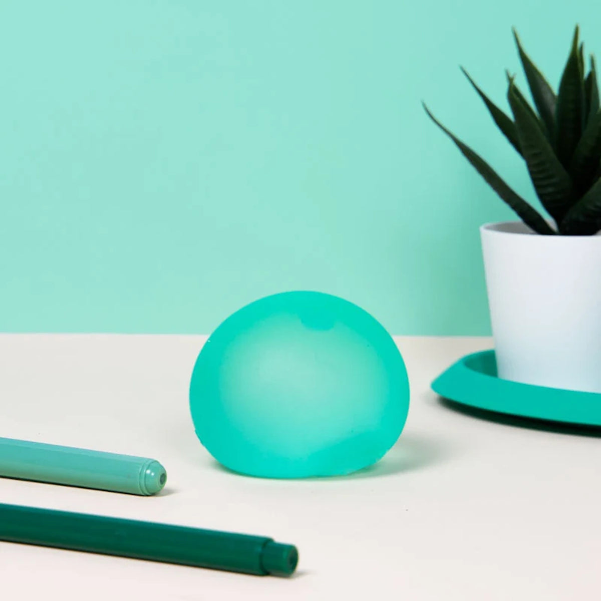 A green squishy stress ball resting on a desk next to green markers and a potted plant.