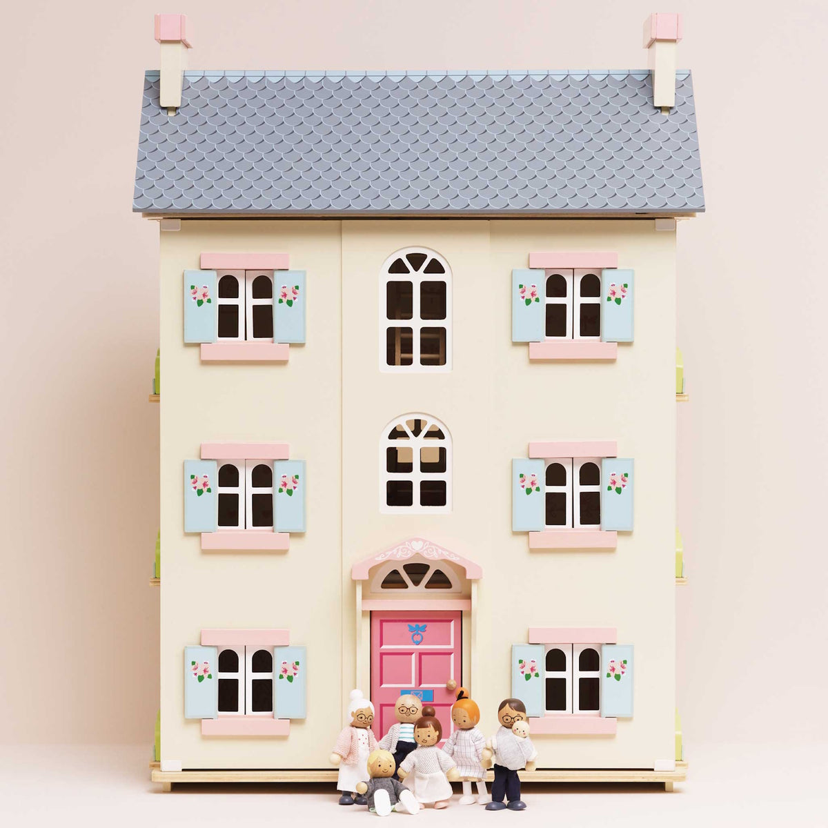 Seven wooden family figures standing in front of a light yellow dollhouse with pink doors and shutters.