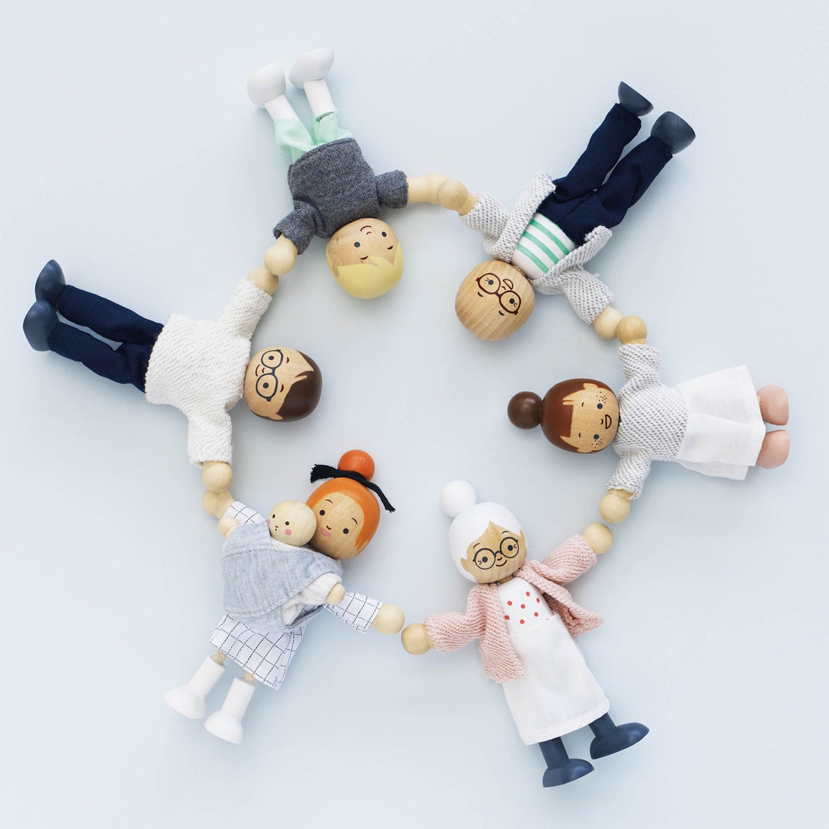 Seven wooden dollhouse family figures lying in a circle holding hands against a light blue background.