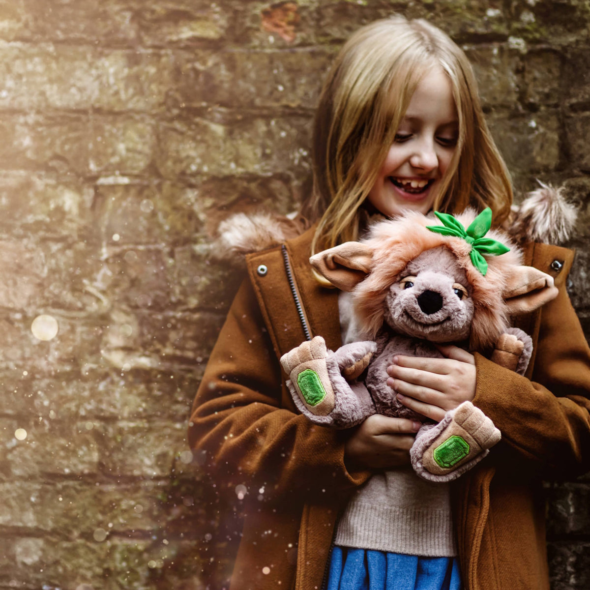 Smiling young girl in a brown coat holding a plush grey troll toy against a stone wall background.