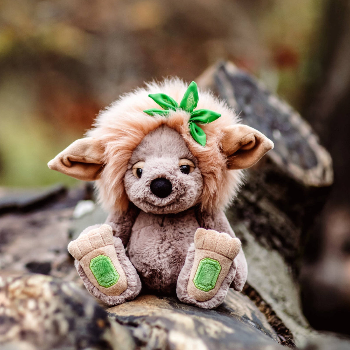 Plush grey troll toy with a fluffy tan mane and green leaf resting on a tree log outdoors.