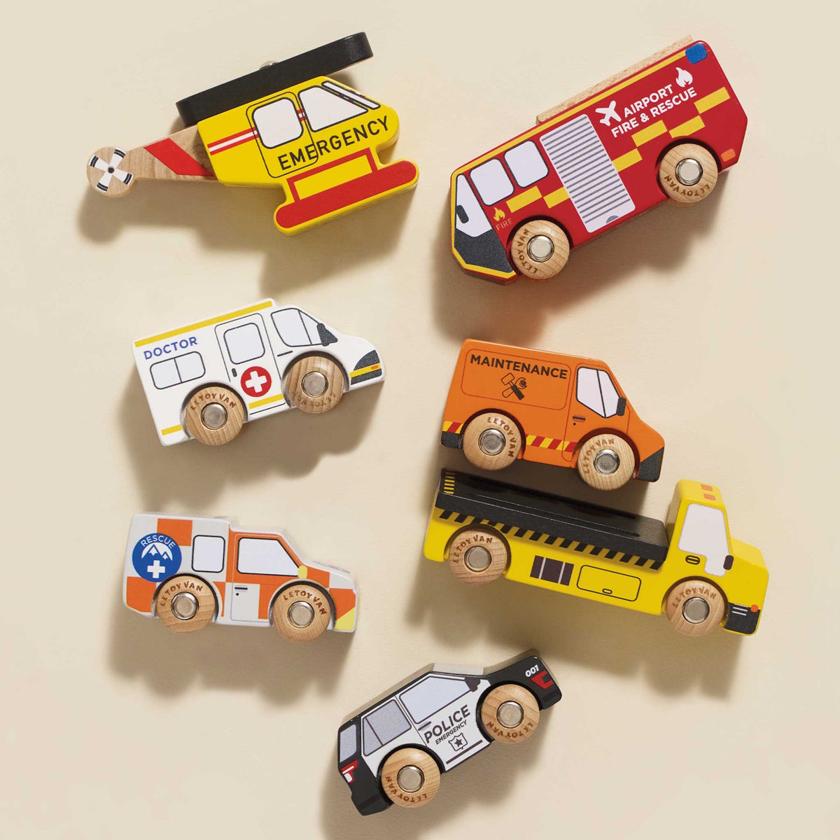 Overhead view of a seven-piece wooden toy emergency vehicle set arranged on a beige background.
