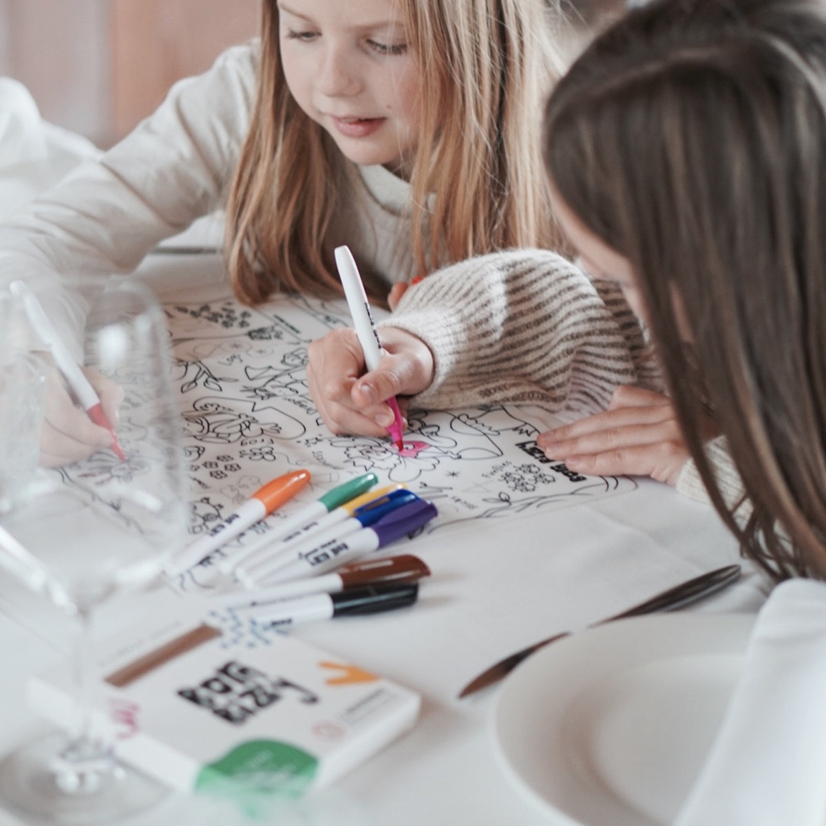 Two girls coloring a large activity mat with markers at a dining table.