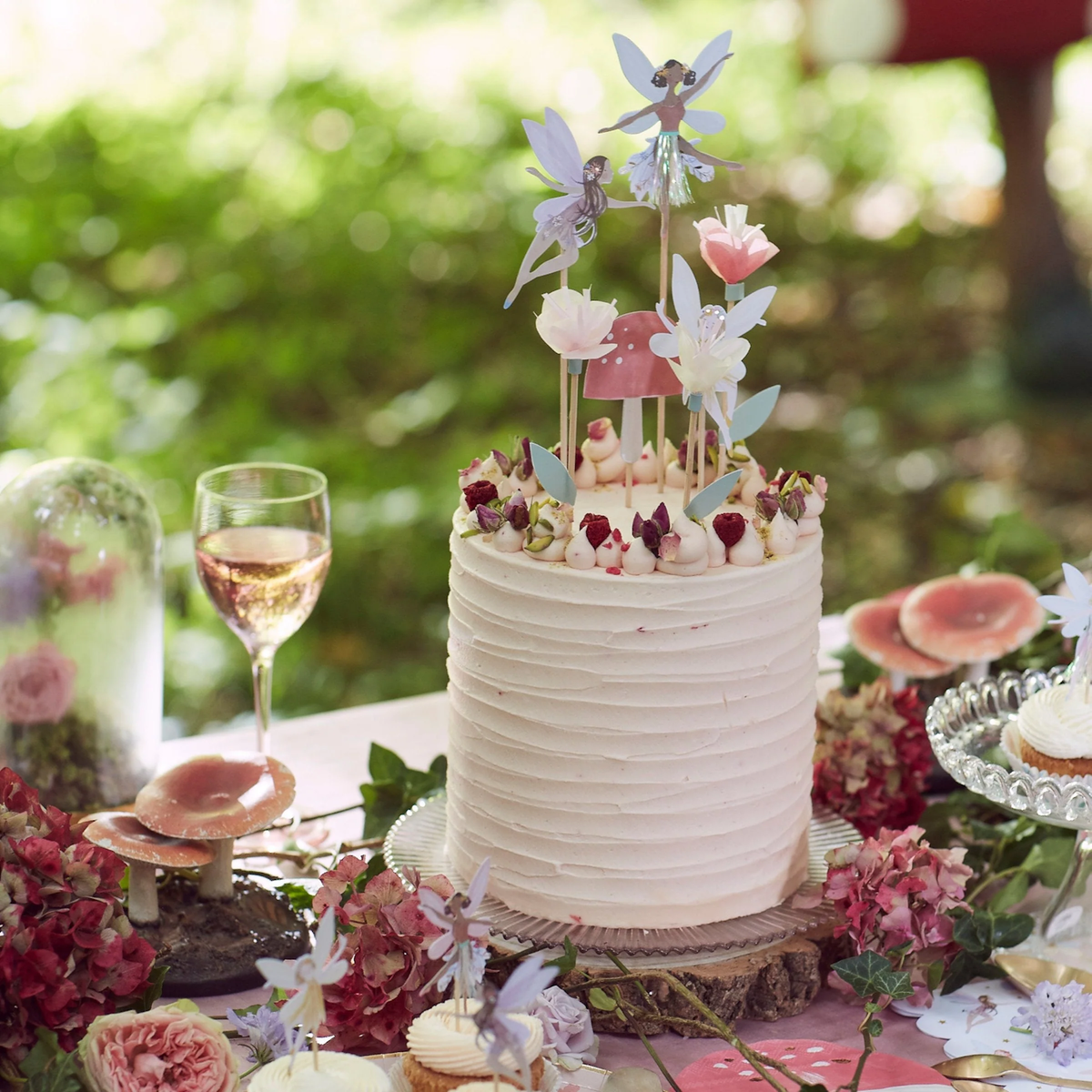 A decorated frosted cake topped with paper fairy toppers, tissue paper flowers, and a pink toadstool on wooden picks, set on an outdoor table.
