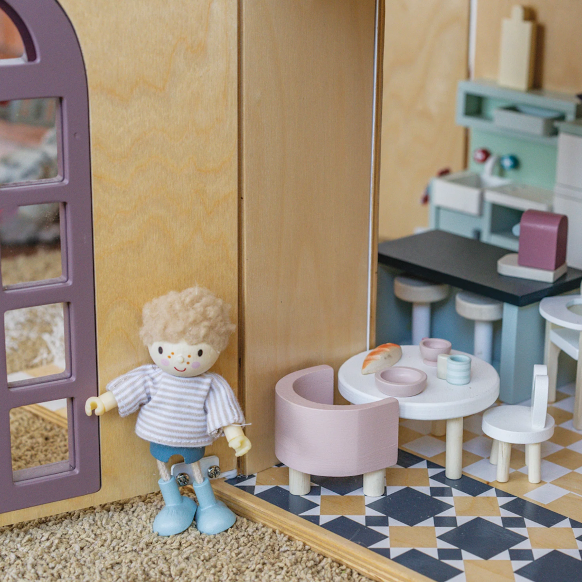 A wooden toy figure stands beside a dollhouse dining room set with a small round table.