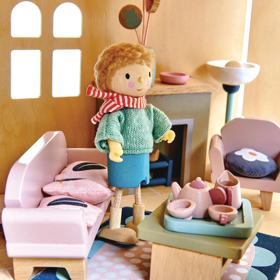 A wooden doll stands in a cozy dollhouse living room near a pink sofa and a tea set.