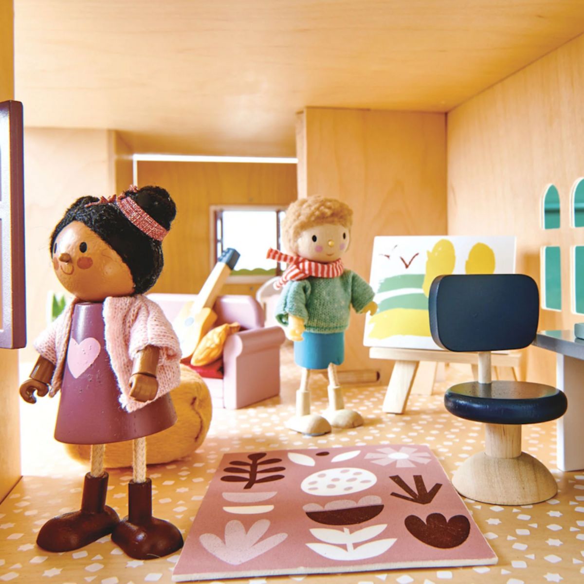 Two wooden dolls stand in a dollhouse room with an easel, office chair, and colorful rug.