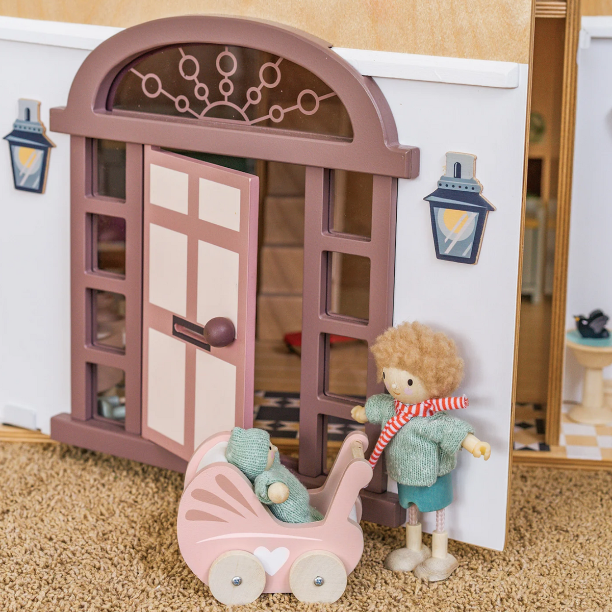 A small toy figure pushes a pink pram with a baby figure in front of an open dollhouse front door.