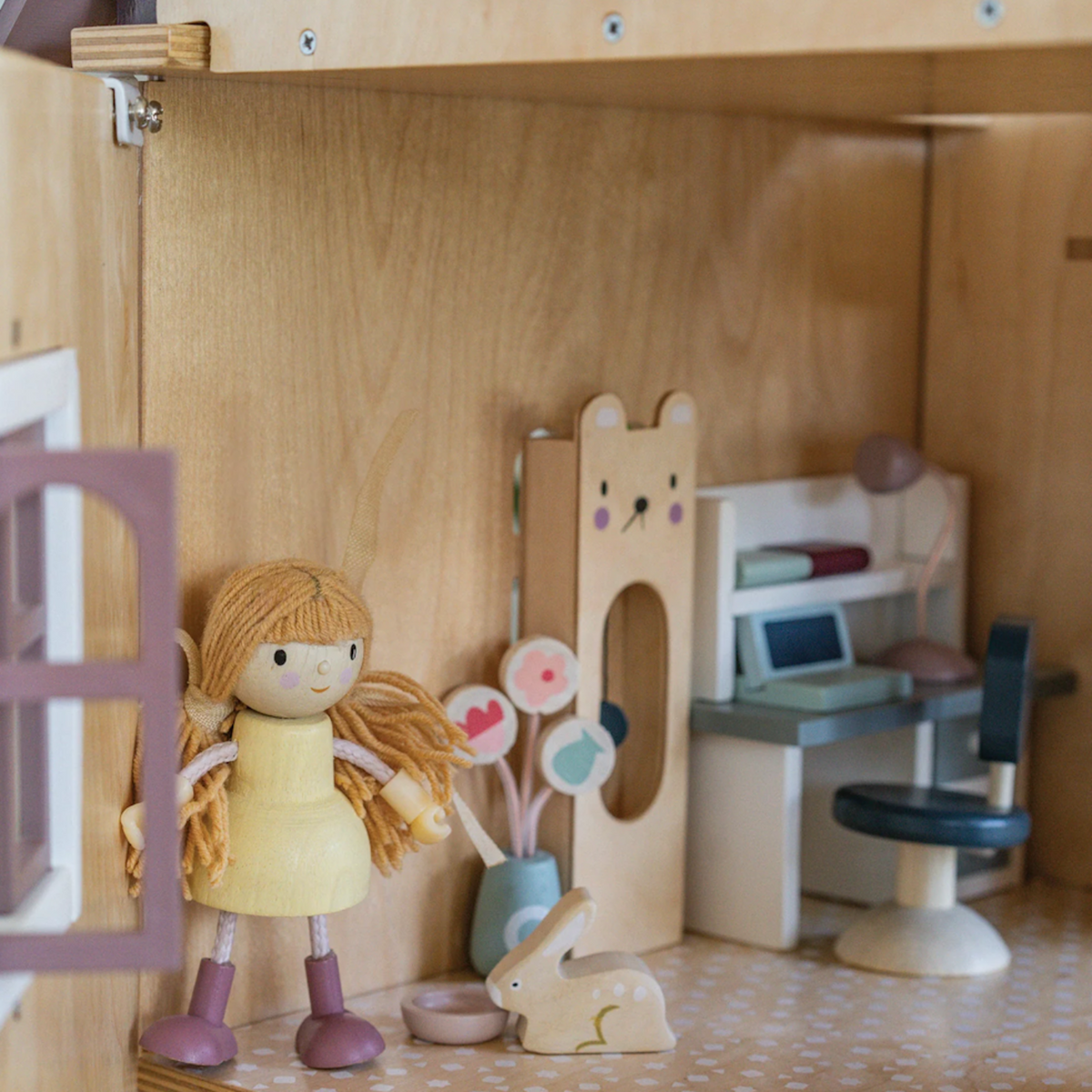 A wooden doll in a yellow dress stands in a miniature dollhouse room with a desk and accessories.