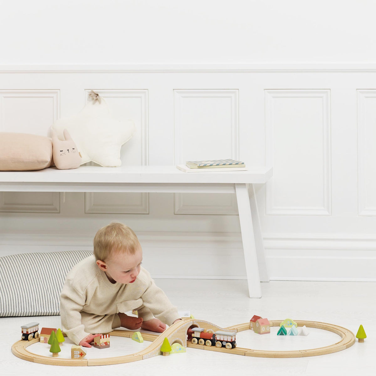 Young toddler playing with a wooden train set on the floor in a brightly lit room.