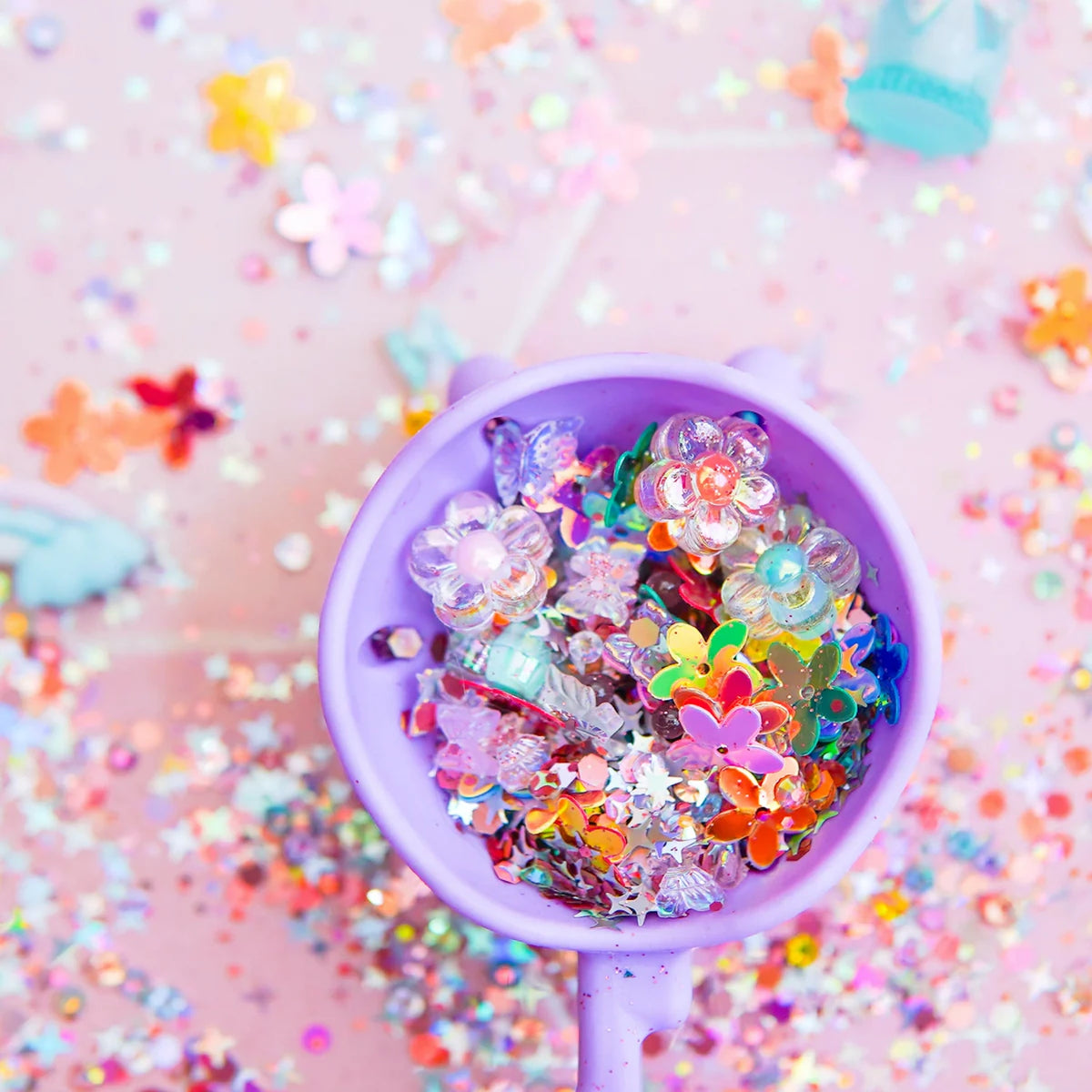 A purple cup filled with fairy-themed trinkets, flowers, and glitter against a sparkly pink background.