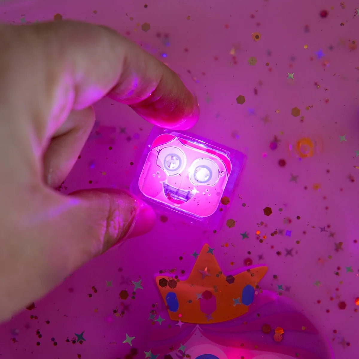 Hand holding a glowing pink cube toy with a smiling face in pink water with glitter.