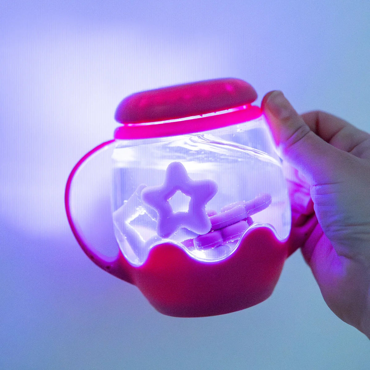 Hand holding a glowing pink Glo Pals Mini Sensory Play Jar filled with water and floating star shapes.