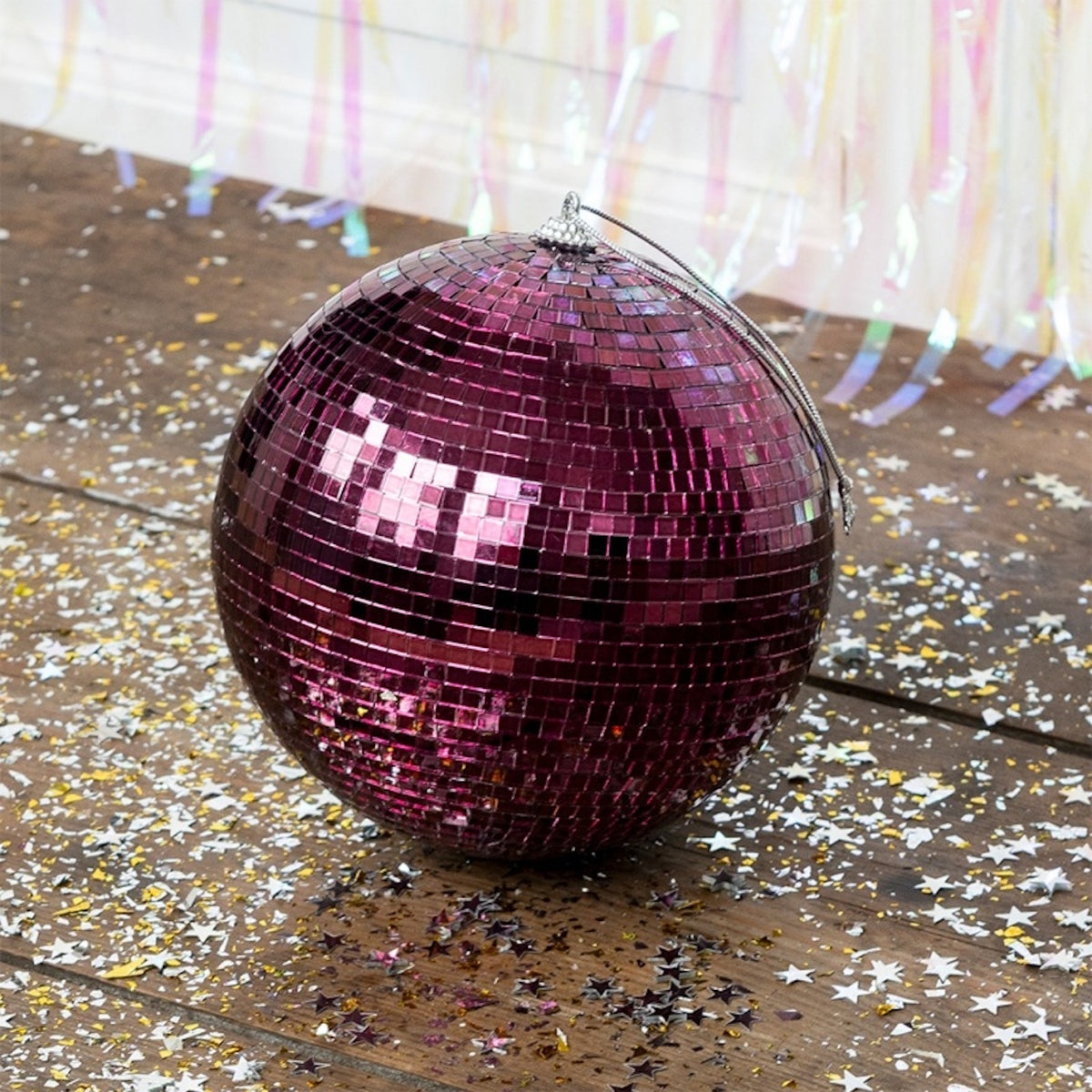 A plum purple disco ball with a silver hanging loop sits on a wooden floor covered in scattered star confetti.