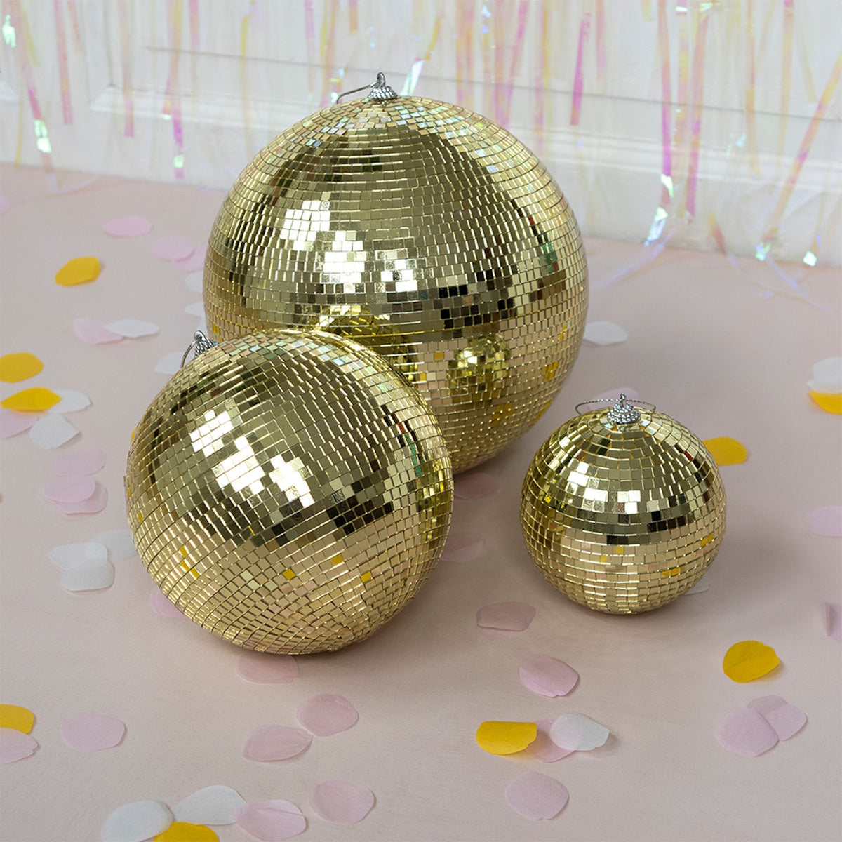 Three shimmering gold disco balls of different sizes are arranged on a light pink surface scattered with yellow, white, and pink confetti.