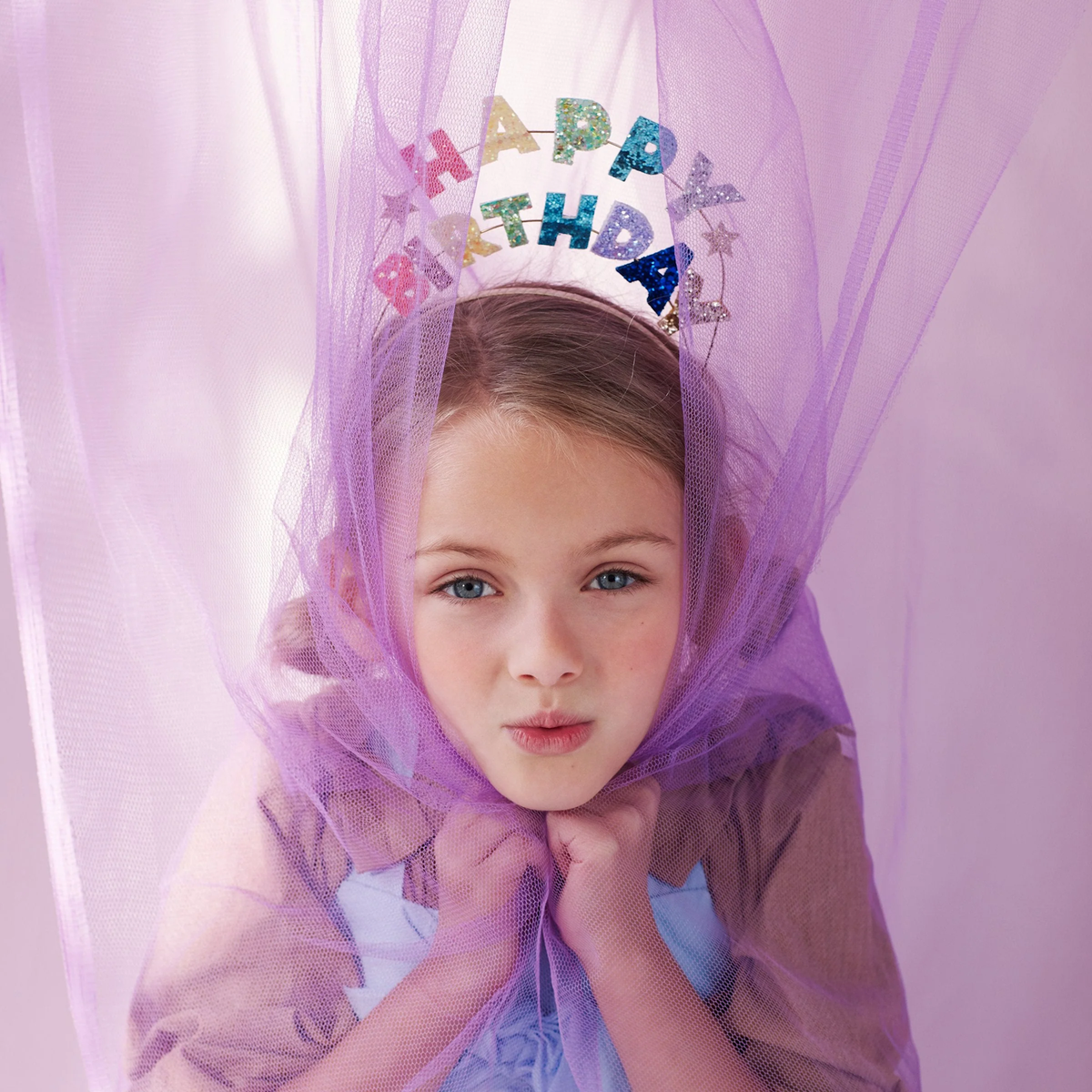 Young girl in a purple tulle veil wears a glittery “HAPPY BIRTHDAY” crown, posing with hands under her chin in a soft, playful portrait.