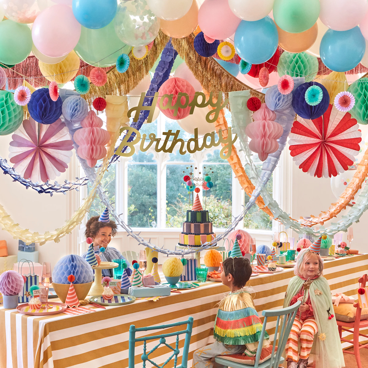 Festive party scene featuring children at a decorated table below a colorful honeycomb Happy Birthday garland.