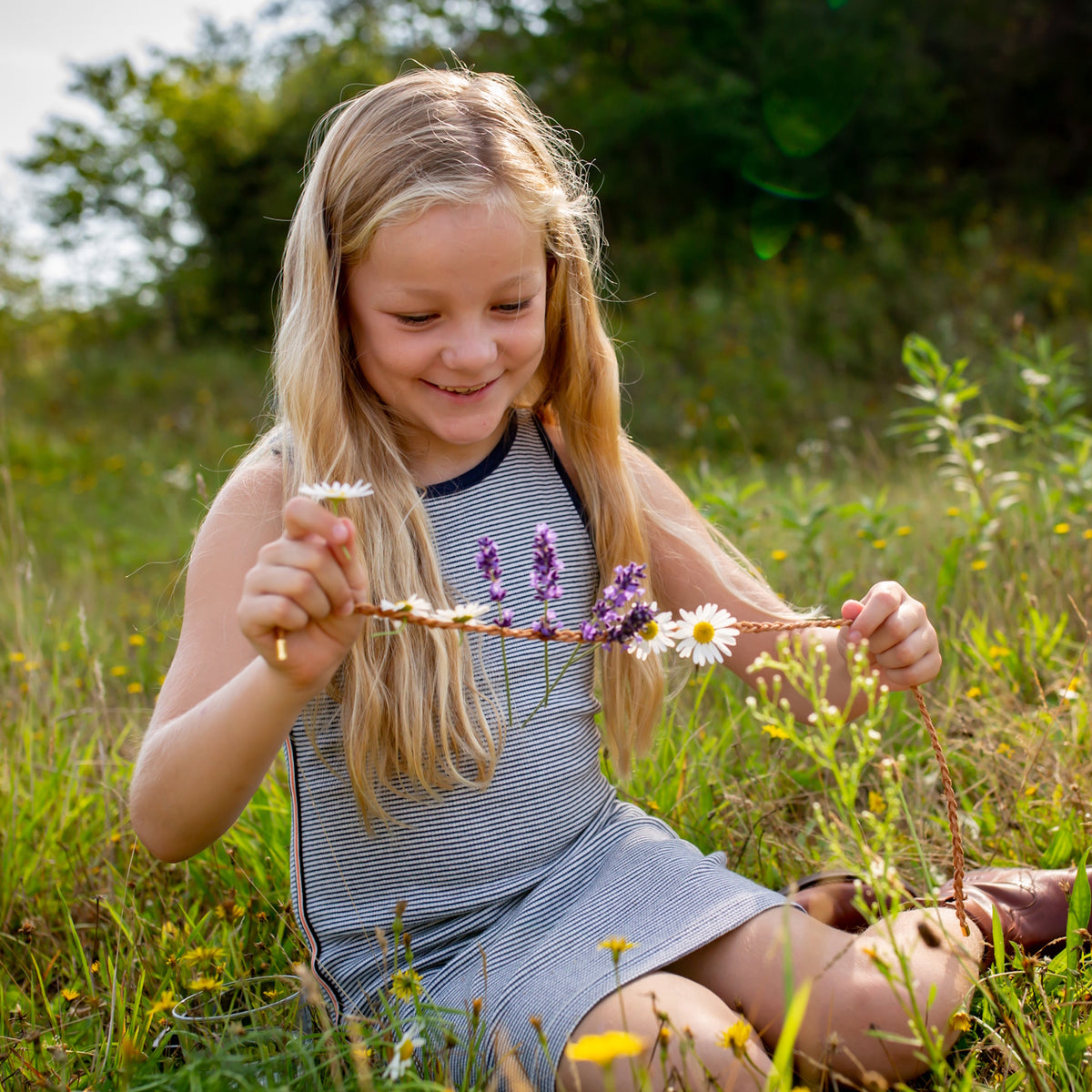 Young girl in a meadow smiling while crafting a DIY necklace with fresh daisies and lavender on a braided cord.