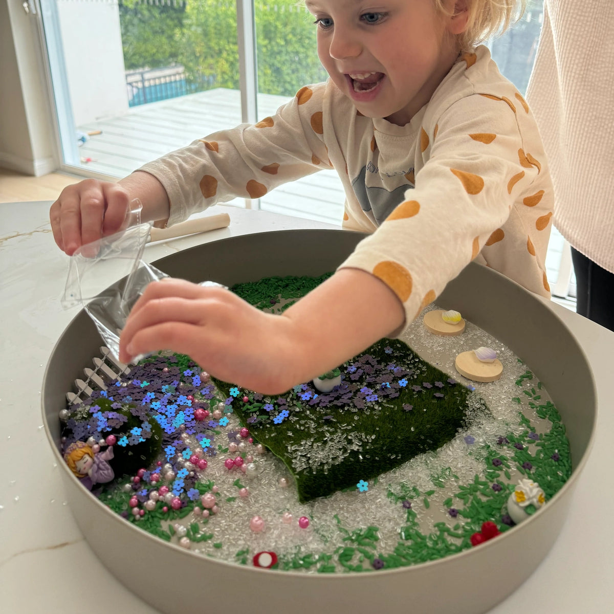 Young child excitedly pouring sequence into a fairy garden sensory tray with moss and floral accessories.