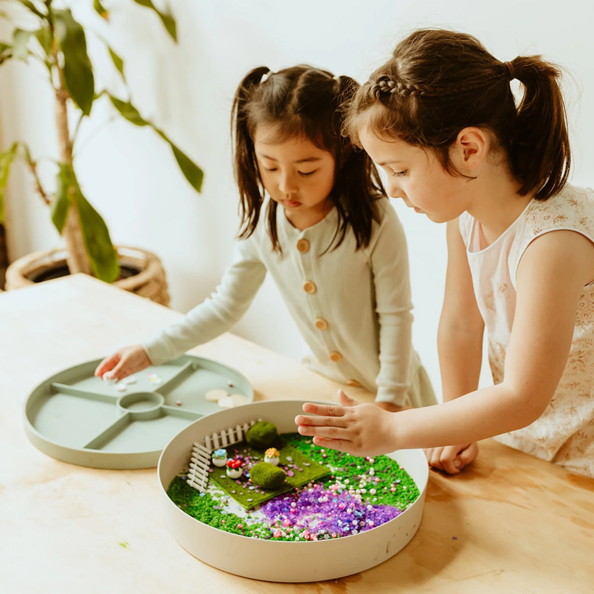 Two young girls playing with a fairy garden sensory tray set up with faux grass, moss, and mini fence pieces.